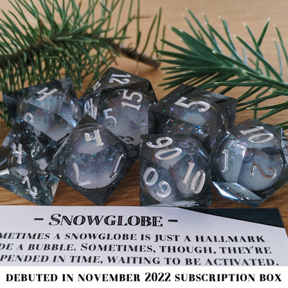 Snow Globe is a 7-piece, translucent grey-blue, sharp edge resin dice set with a liquid core of snow white glitter, inked in bright silver.